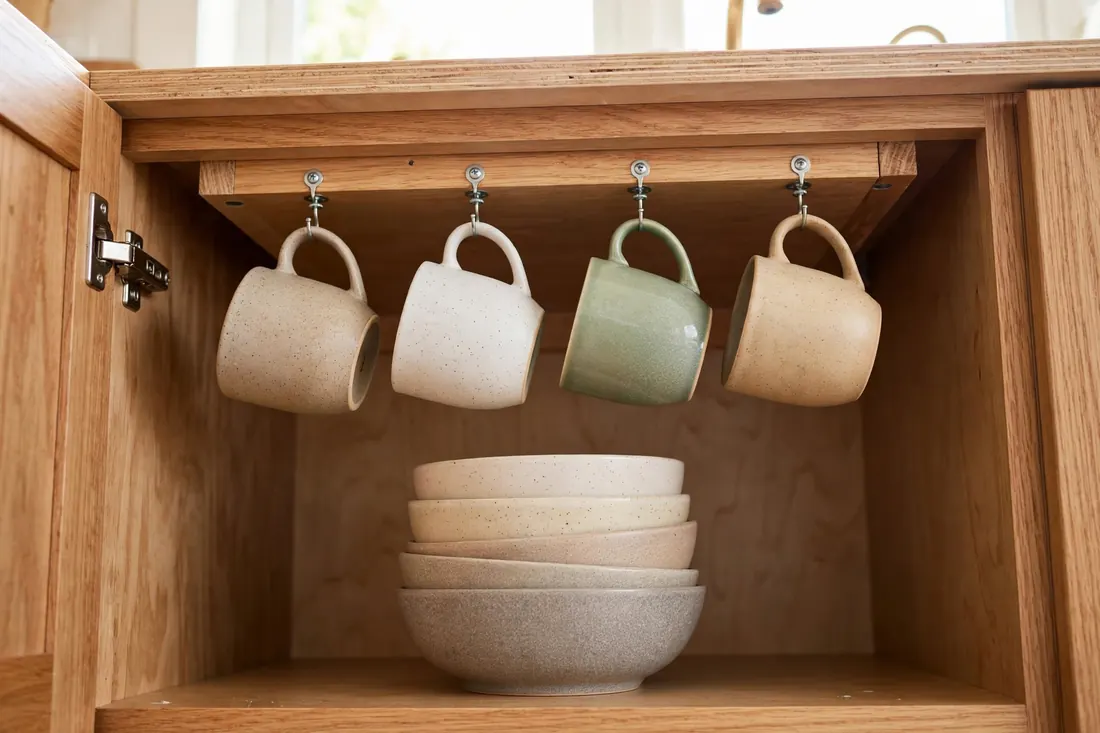 Coffee mugs hanging from hooks under a cabinet shelf above stacked bowls