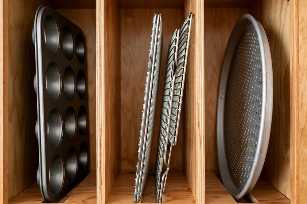 Muffin tins, wire cooling racks, and a round pizza pan filed upright in a cabinet divider