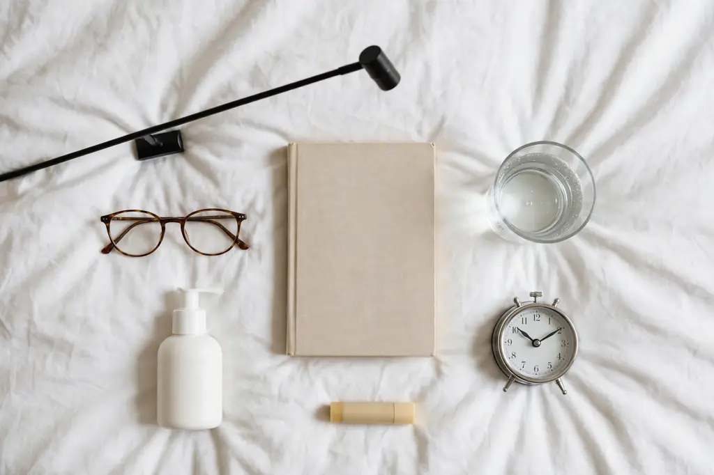 Nightstand essentials arranged on a bed: lamp, glasses, book, water, lip balm, lotion, alarm clock