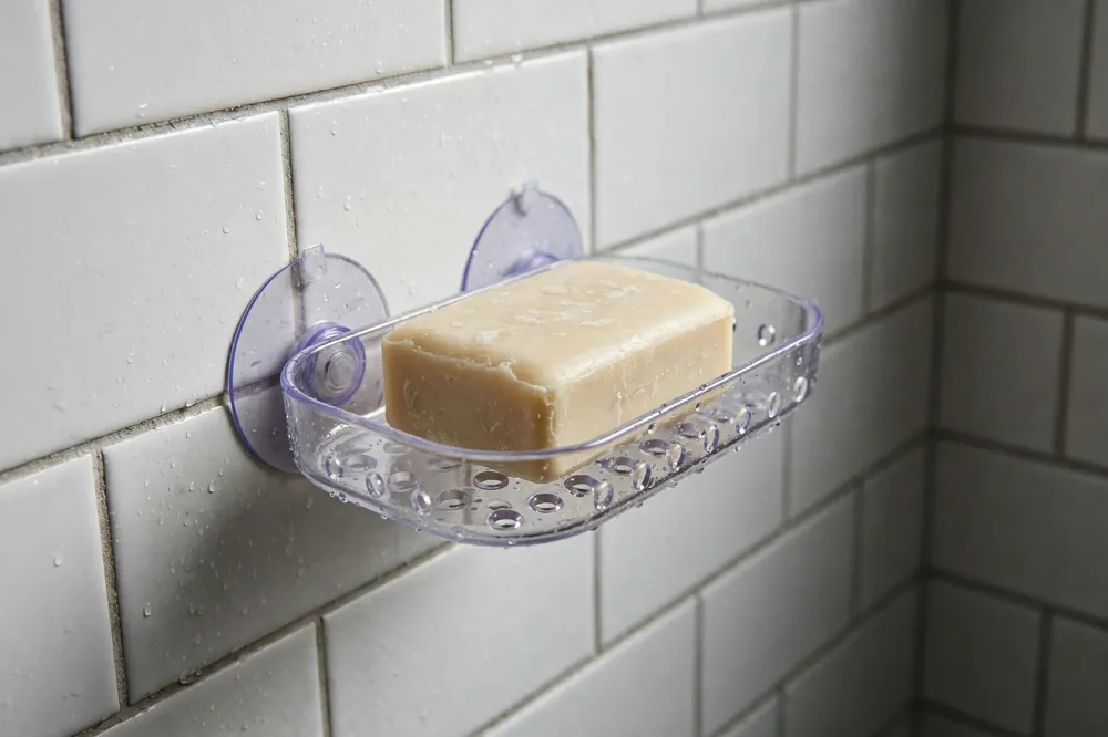 Suction soap dish with drainage holes holding a bar of soap in a tiled shower