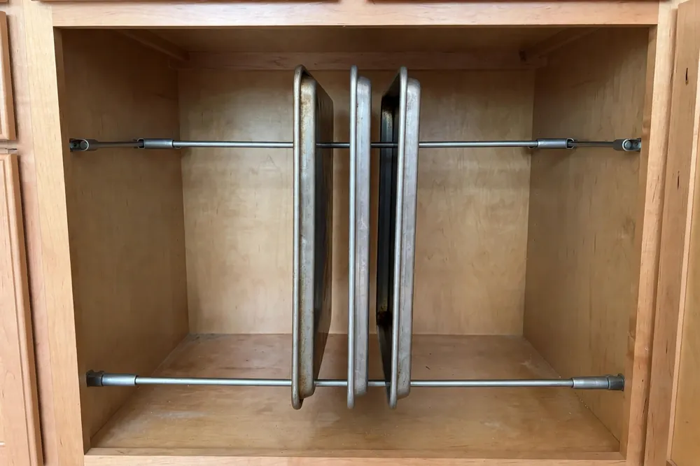 Metal tension rods inside a kitchen cabinet holding baking sheets upright between them